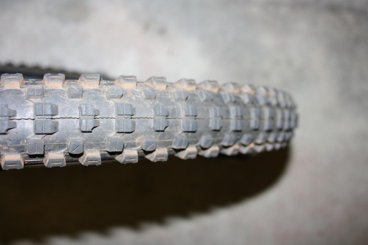 Kenda Nevegal: Close-up image of a mountain bike tire showing its rugged tread pattern. The tire is slightly dirty, indicating use on uneven terrain. The background is blurred, emphasizing the tire's details.