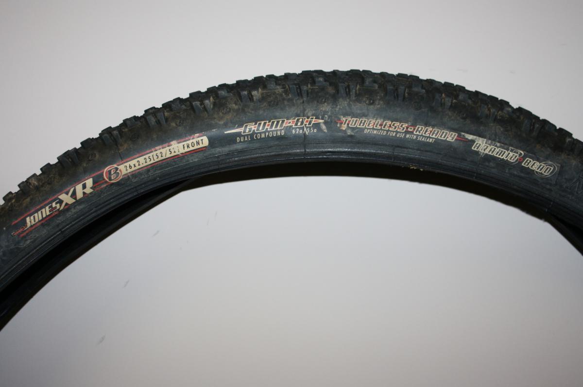 Bontrager Jones XR: A close-up image of a mountain bike tire showing the tread pattern, with labels indicating the model "Jones XR" and specifications including size (26 x 2.25) and features such as "tubeless ready" and "dual compound." The tire appears slightly dirty, suggesting off-road use.