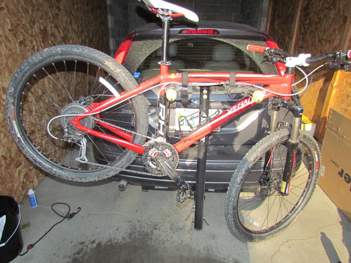 Specialized Rockhopper Comp Disc: A red mountain bike hung upside down on a bike rack in a garage, with dirt on its tires. A dark-colored vehicle can be seen in the background, and there are some tools and equipment on the ground. The garage walls are made of wood panels.