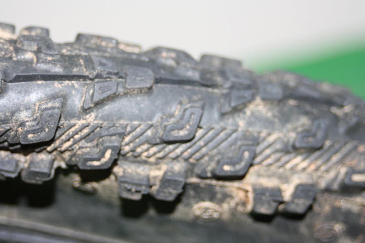 Bontrager Jones XR: Close-up view of a textured tire tread, showcasing its intricate patterns and grooves, with some dirt visible on the surface. The background features a blurred green and white surface.