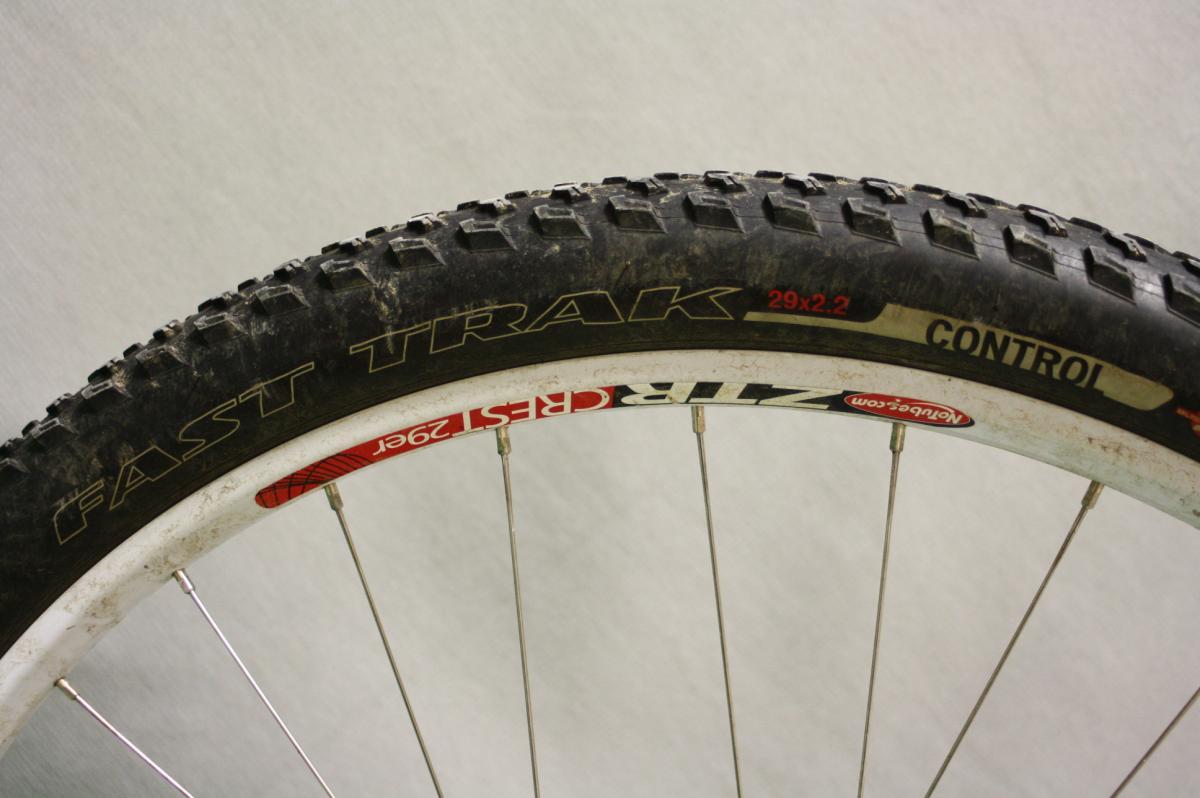 Specialized Fast Trak: Close-up of a mountain bike tire mounted on a wheel, featuring the brand name "FAST TRAK" and specifications "29x2.2." The tire has a rugged tread pattern suitable for off-road terrain, and the wheel rim is white with a red emblem labeled "Crest." The background is a neutral gray.