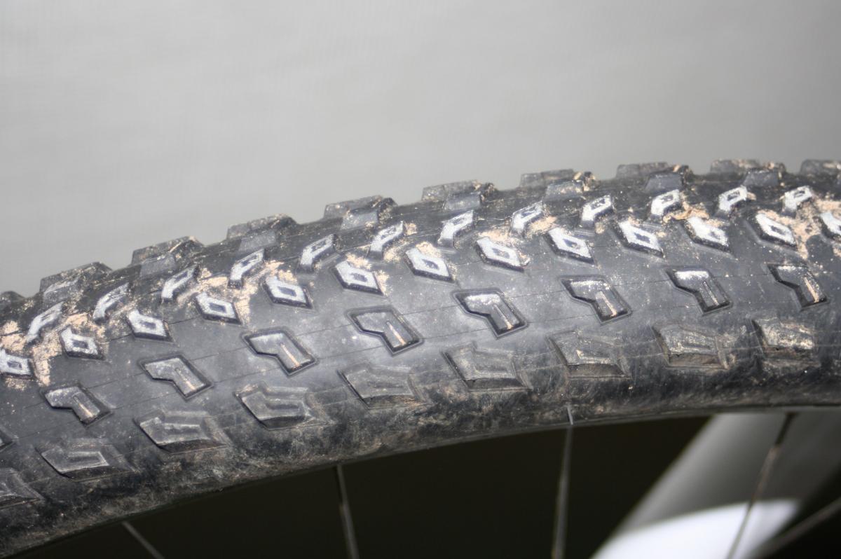 Specialized Fast Trak: Close-up of a bicycle tire, showcasing the textured tread pattern and some dirt, set against a light-colored background. The image highlights the details of the rubber surface and the design intended for traction.