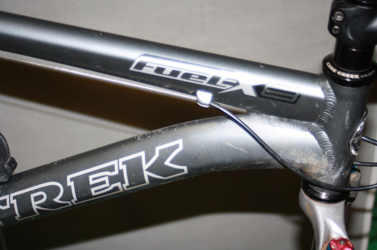 Trek Fuel EX 8: Close-up of a Trek bicycle frame, showcasing the "Fuel EX" model designation and some signs of wear, including scratches. The focus is on the frame's design and details, including the welds and cable routing.