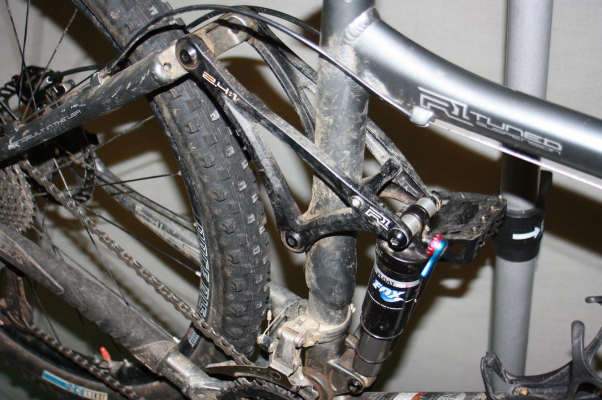 Trek Fuel EX 8: Close-up of a mountain bike's rear suspension system, showcasing the shock absorber and linkage mechanism. The bike frame is covered with dirt, indicating off-road use, and features a prominent tire with rugged tread.