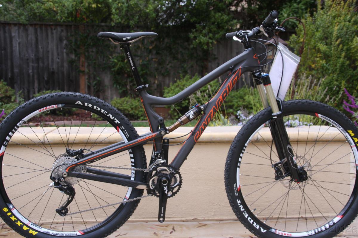 Santa Cruz Tallboy LT: A modern mountain bike with a sleek black and orange frame, featuring large knobby tires designed for off-road use. The bike is shown from the side, highlighting its suspension system and gear mechanism. The background includes greenery, suggesting an outdoor setting.