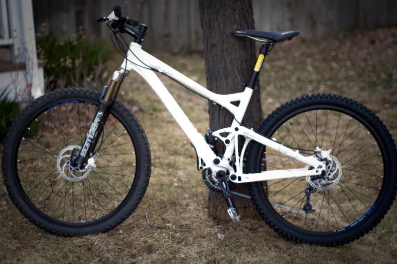 Giant Trance 1: A white mountain bike with thick tires and a dual suspension system, leaning against a tree in a grassy area. The bike features a front shock absorber and a black seat, showcasing a rugged design suitable for off-road cycling.