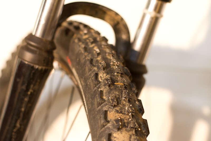 Specialized Resolution Sport: Close-up view of a muddy mountain bike tire, showcasing its textured tread pattern and a portion of the bike's fork in the background, emphasizing the rugged design suitable for off-road terrain.