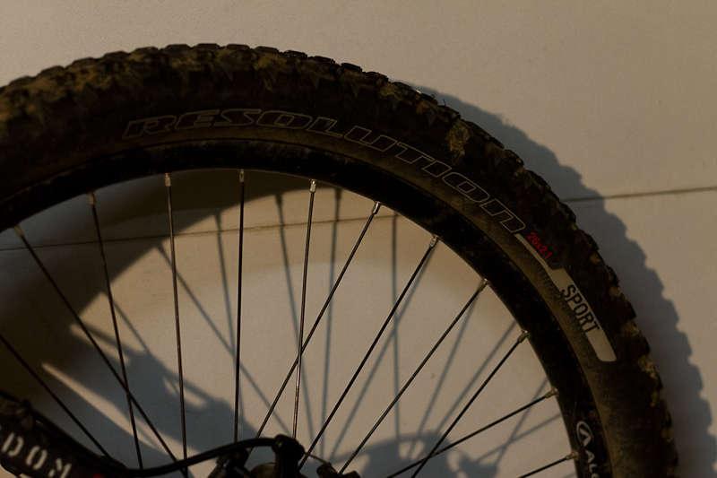 Specialized Resolution Sport: Close-up image of a bicycle tire resting against a light background, featuring the tread pattern and markings that read "RESOLUTION" and "Sport." The tire appears slightly muddy, indicating recent use.