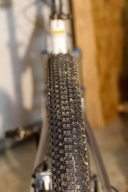 Kenda Small Block Eight: Close-up view of a mountain bike tire, showcasing its tread pattern and some dirt, with the bike frame partially visible in the background. The setting appears to be an indoor space, possibly a garage.