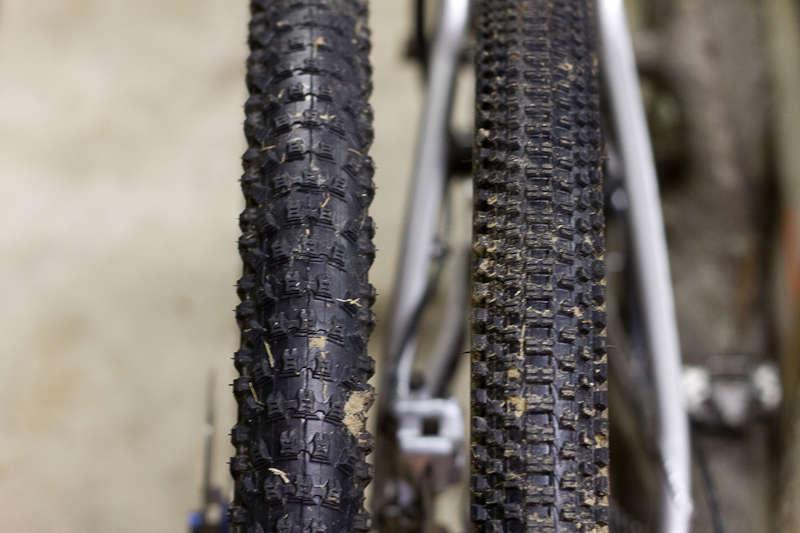 Kenda Slant Six: Close-up image of two bicycle tires side by side, showcasing their different tread patterns. The left tire features a more aggressive, knobby design suitable for off-road biking, while the right tire has a smoother, less pronounced tread ideal for road use. The background is blurred, highlighting the tires' textures and details.