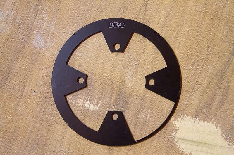 BBG Bashguard: Black circular metal part with four cutouts and the letters "BBG" engraved on top, placed on a wooden surface.
