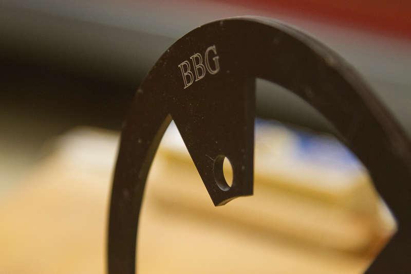 BBG Bashguard: Close-up of a black metal ring with an engraved "BBG" logo, featuring a cutout and a small hole. The background is blurred, suggesting a workshop or tool environment.