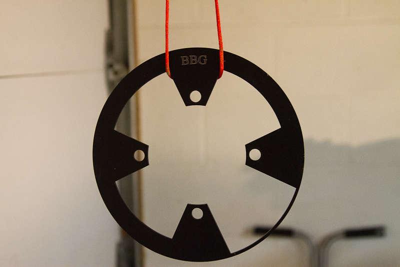 BBG Bashguard: Black circular target with cutouts, hanging by a red string against a neutral background. The target features the letters "BBG" engraved at the top.