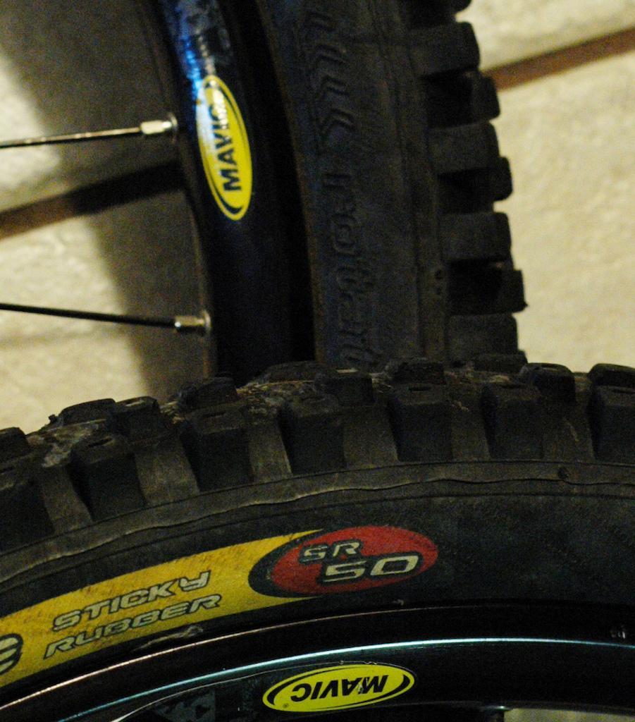 ITS Fro Lite DH: Close-up image of two bicycle tires, one positioned above the other. The top tire features a visible tread pattern, with a brand logo partially obscured. The bottom tire displays markings indicating it is made of "sticky rubber" and has a label reading "SR 50." The background is a simple, neutral surface, highlighting the tires.