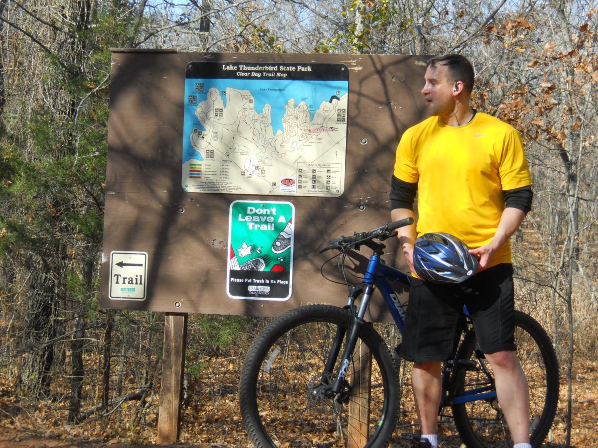 Diamondback Overdrive Sport: A person standing next to a trail map sign at Lake Thunderbird State Park, wearing a yellow shirt and black shorts, holding a bike helmet and standing beside a mountain bike. In the background, there are trees with autumn foliage.