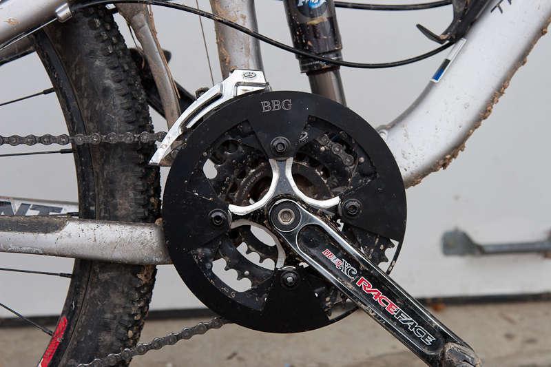 BBG Bashguard: A close-up view of a bicycle's crankset and chainrings, featuring a black chainring with the label "BBG" and a silver crank arm marked "Race Face." The bicycle appears to be used, with visible dirt and mud on the components.