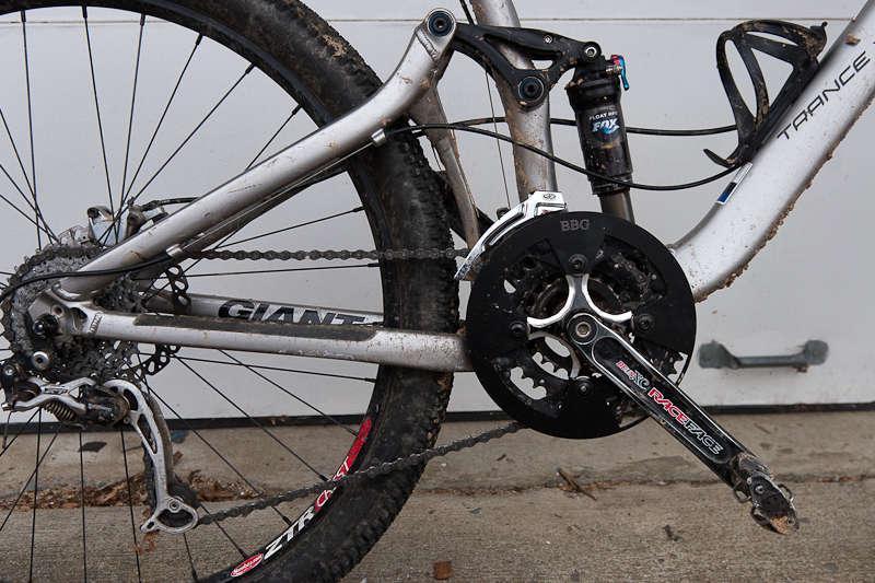 BBG Bashguard: A close-up view of a mountain bike's rear drivetrain, featuring a dirty silver frame with the name "Giant" visible. The photo highlights the black crankset, chainguide, and rear derailleur, with visible wear marks on the components. The background is a concrete surface, suggesting outdoor conditions.
