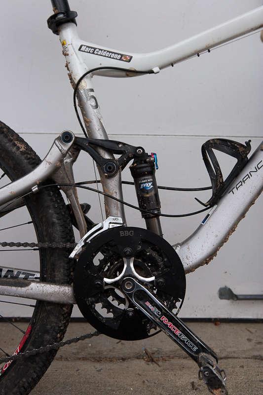 BBG Bashguard: Close-up of a mountain bike frame showcasing the drivetrain, including the crankset and derailleur. The frame is slightly muddy, indicating recent use in off-road conditions. A shock absorber is visible near the rear suspension, and the bike features brand markings such as "Trance" and "Race Face."