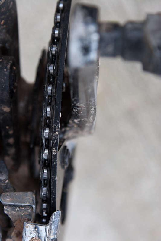 BBG Bashguard: Close-up view of a bicycle chain and gear mechanism, showing details of the chain links and the gear teeth, with a slightly dirty appearance indicating usage.