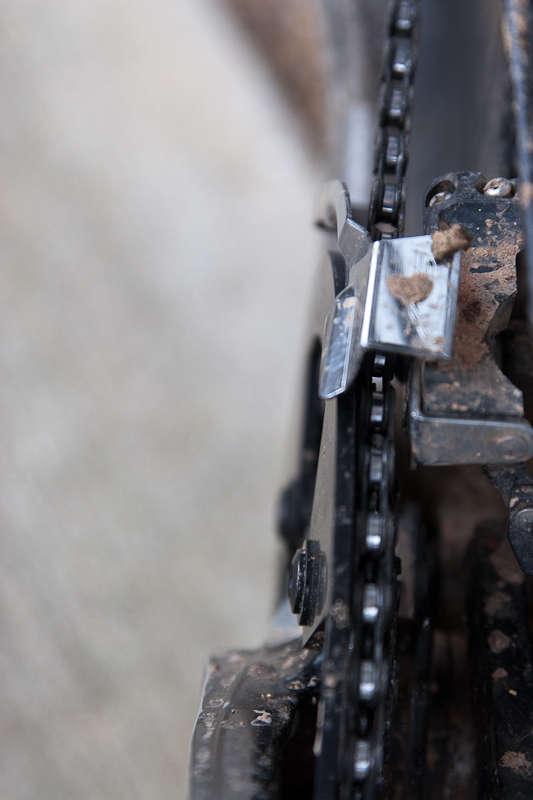 BBG Bashguard: Close-up view of a bicycle chain and derailleur, showing dirt and grime on the components. The image highlights the mechanical details of the chain's tension system and the surrounding areas.