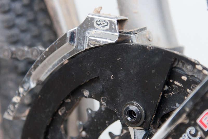 BBG Bashguard: Close-up of a bicycle's front derailleur and chainring, showcasing the detailed mechanism and components. The image highlights dirt and wear, indicating use in various terrains.