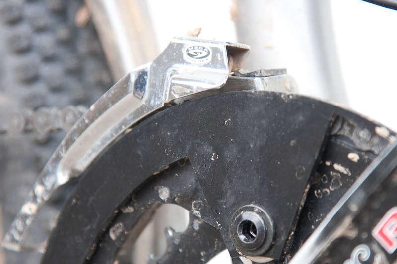BBG Bashguard: Close-up image of a bicycle's derailleur and chainring, showcasing details such as dirt accumulation and metal components. The focus is on the mechanism used for shifting gears, highlighting its design and functionality.