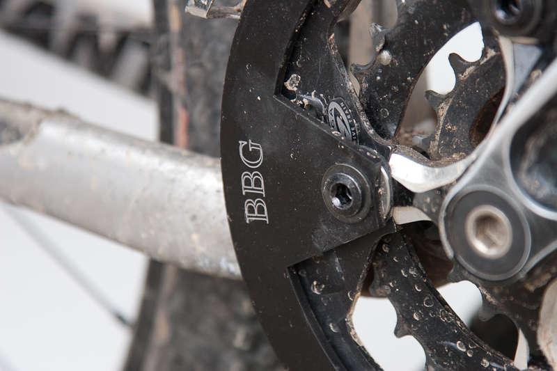 BBG Bashguard: Close-up image of a bicycle's drivetrain components, featuring a black chainring with the engraving "BBG," showing signs of dirt and wear, attached to a metallic frame. The photograph highlights the intricate details of the gear and chain setup.