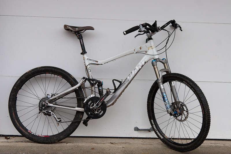 Giant Trance X2: A white and silver mountain bike leaning against a gray garage wall. The bike features thick tires, front suspension, and a brown seat.
