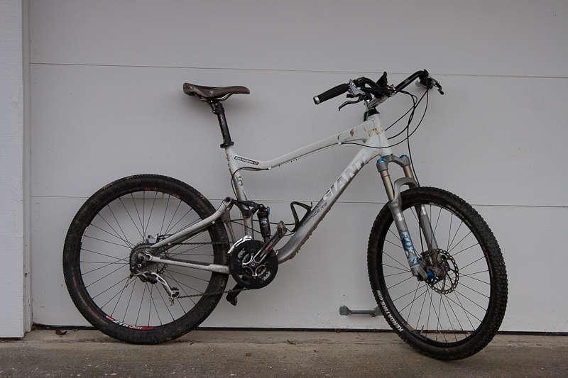 Giant Trance X2: A silver mountain bike leaning against a white wall, featuring a brown saddle, black tires with visible tread, and front suspension forks. The bike is equipped with disc brakes and a rear derailleur, showcasing a rugged design suitable for off-road cycling.