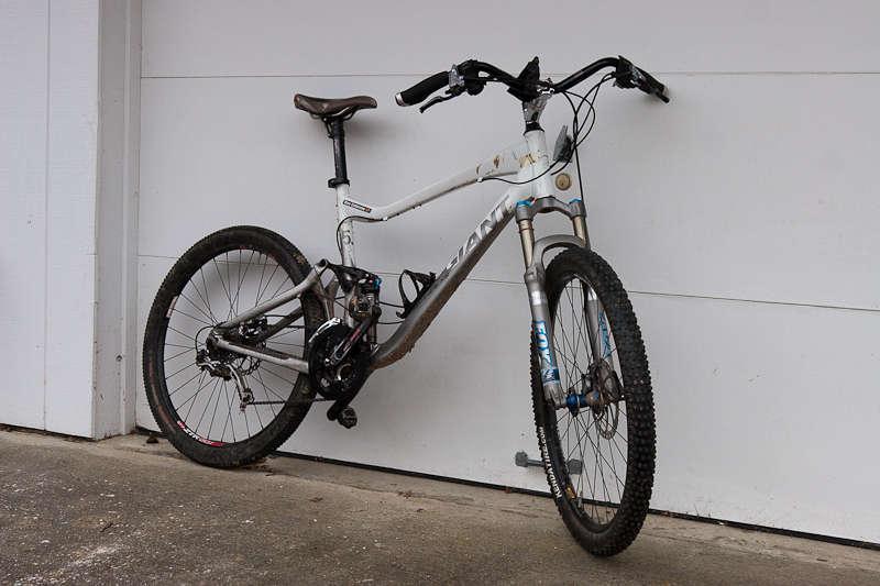 Giant Trance X2: A mountain bike leaning against a white garage door, featuring a gray and white frame, thick tires, and visible gears.