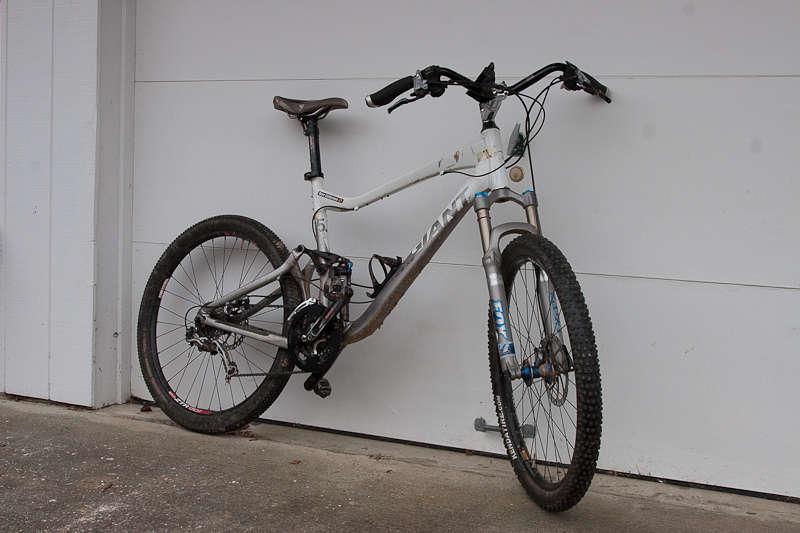 Giant Trance X2: A mountain bike leaning against a garage wall, featuring a silver and gray frame, thick tires with knobby tread, and disc brakes. The bike has a visible gear system and a comfortable saddle, suggesting it's designed for off-road riding.