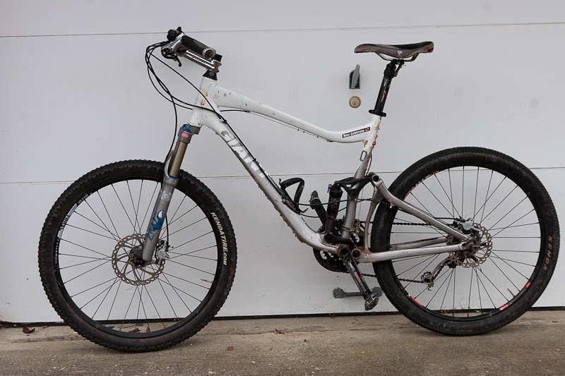 Giant Trance X2: A mountain bike with a silver and white frame, featuring a front suspension fork and wide tires, is leaning against a garage door. The bike has disc brakes and a gear shifter attached to the handlebars.