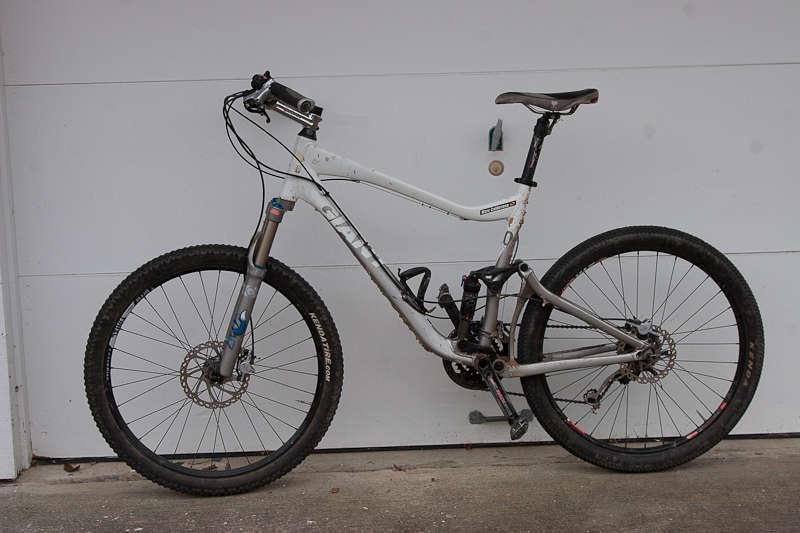 Giant Trance X2: Mountain bike leaning against a garage door, featuring a silver frame, front suspension, and knobby tires suitable for rugged terrain.