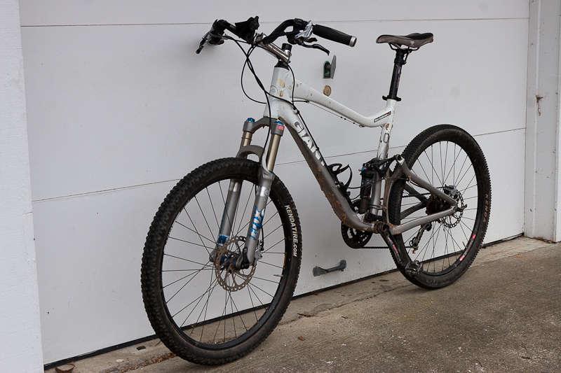 Giant Trance X2: Mountain bike leaning against a garage door, featuring a white frame, front suspension, large knobby tires, and a dirty appearance, suggesting recent use.