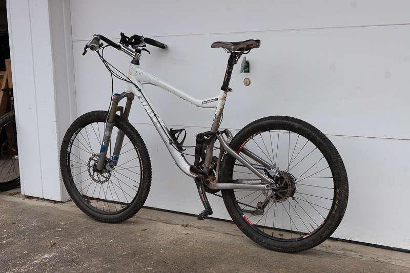 Giant Trance X2: A white mountain bike with a dual suspension, positioned against a garage door. The bike features disc brakes, knobby tires, and a small water bottle holder attached to the frame. The environment appears to be a residential garage setting.
