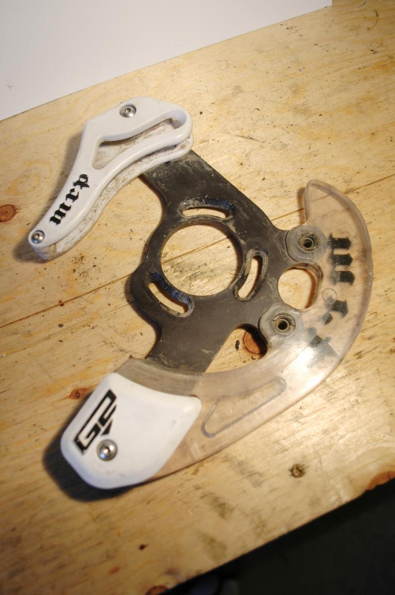 MRP G2: Image of a bicycle chain guide resting on a wooden surface. The guide features a metal base with a transparent plastic casing and white plastic components, marked with the brand "mcrp." The overall appearance suggests it has been used, showing signs of wear and dirt.