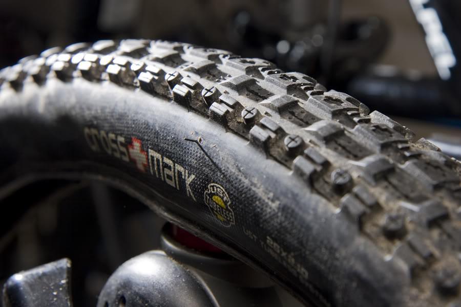 Maxxis Crossmark: Close-up image of a mountain bike tire showcasing its tread pattern. The tire is marked with "CROSSMARK" branding and features small, distinct dots on the surface. The tire shows signs of use with dirt and debris on it, indicating it has been used for off-road cycling. The background is slightly blurred, focusing attention on the tire detail.