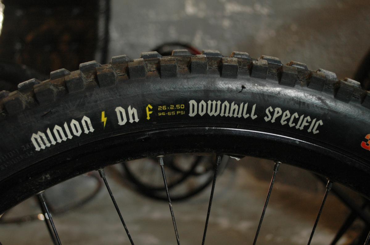 Maxxis Minion DHF: A close-up image of a mountain bike tire featuring the text "Minion DHF 26 x 2.50 Downhill Specific." The tire showcases a rugged tread pattern and is viewed from the side, with background elements suggesting a workshop or garage setting.