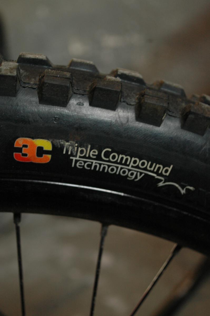 Maxxis Minion DHF: Close-up image of a bicycle tire showcasing the text "3C Triple Compound Technology" printed on the sidewall, emphasizing the tire's advanced design and performance features. The background is slightly blurred, focusing attention on the tire details.