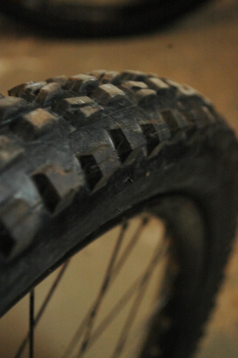 Maxxis Minion DHF: Close-up view of a rugged bicycle tire with a textured tread pattern, designed for traction on various surfaces. The background features blurred outlines of additional bike wheels, suggesting an indoor setting or workshop.