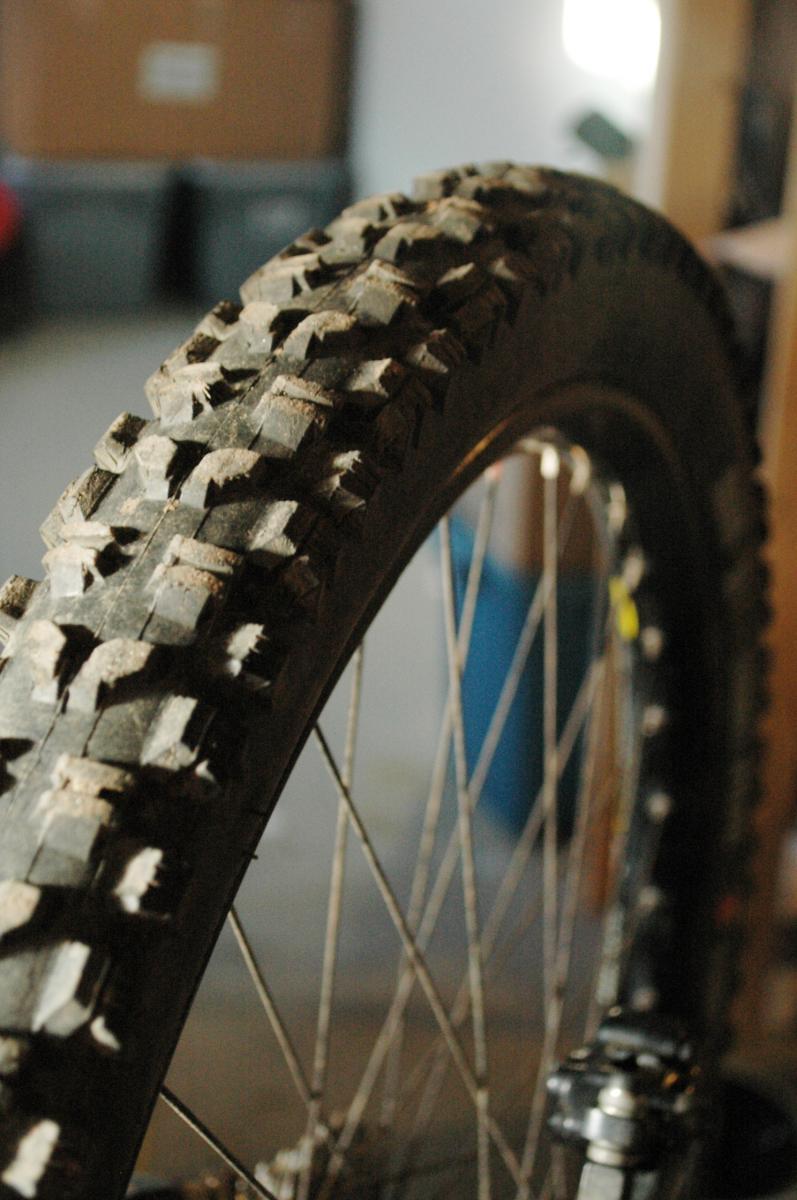 Maxxis Minion DHR: A close-up view of a mountain bike tire with a rugged tread pattern, showcasing dirt on the surface. The background is blurred, with hints of bike components and storage boxes visible.