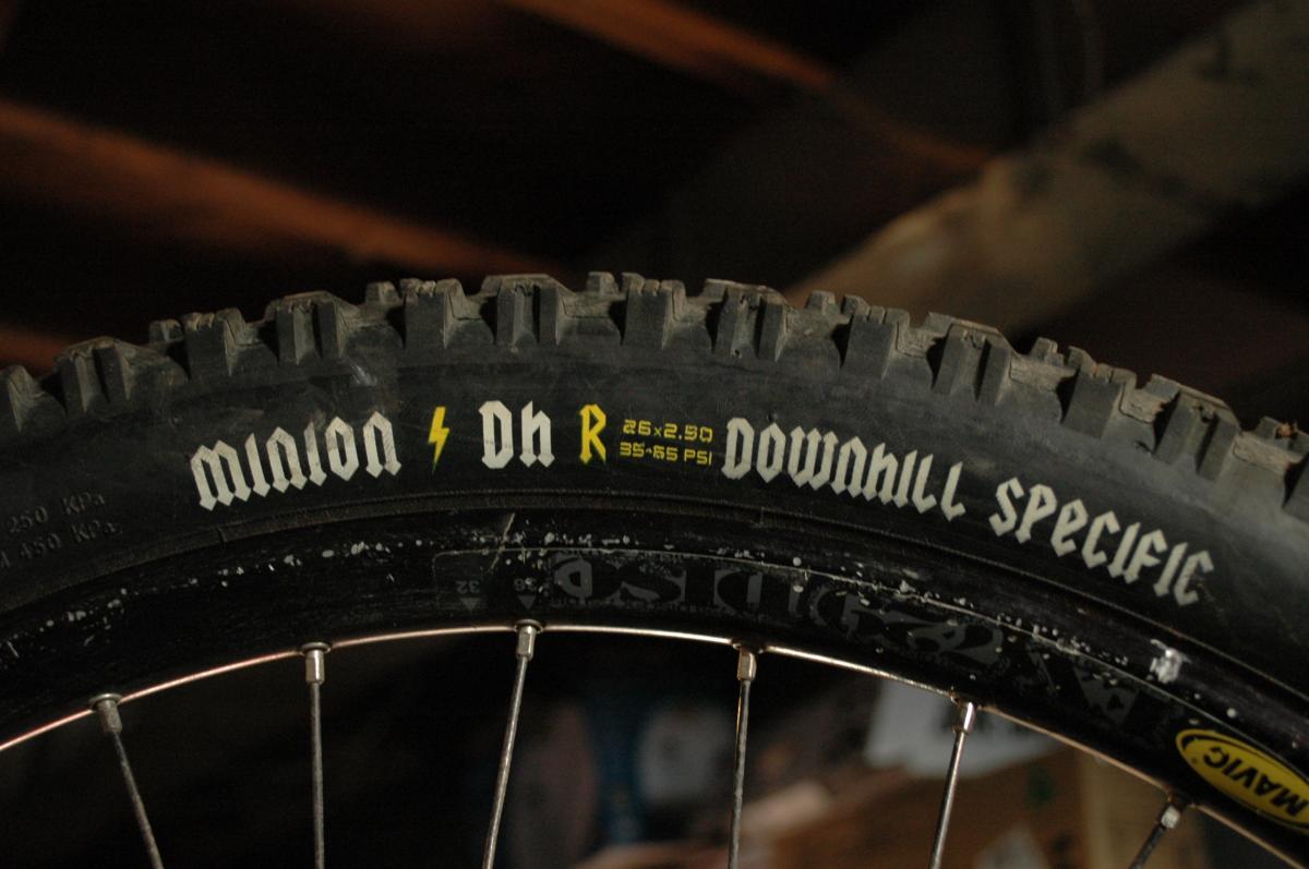 Maxxis Minion DHR: Close-up image of a mountain bike tire displaying the brand name "Minion" with a lightning bolt emblem, and the specification "DH R" alongside the dimensions "26 x 2.50" and recommended pressure range "35-65 PSI." The tire features a tread pattern designed for downhill riding. The background is dimly lit, suggesting a garage or workshop setting.