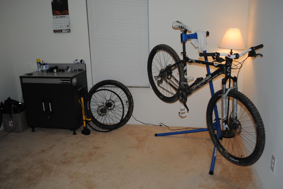 Kenda Nevegal: A mountain bike is mounted on a blue repair stand in a carpeted room, with one wheel lying on the floor nearby. A black cabinet sits next to the bike, and a lamp is illuminated beside it. A calendar displaying December hangs on the wall, with a window featuring blinds behind the bike. Various tools and bike accessories are visible on the cabinet's surface.