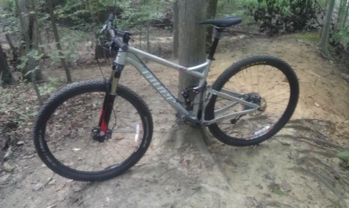 Niner Jet 9: A silver mountain bike leaning against a tree on a dirt trail surrounded by trees and foliage.
