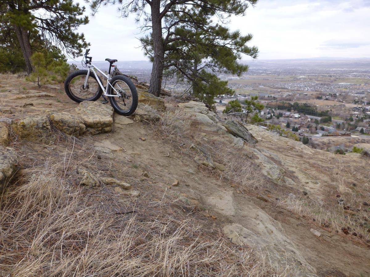 A white mountain bike rests on rocky terrain near the edge of a hill, with a panoramic view of a valley and distant buildings in the background. The landscape features sparse grass and trees, suggesting a rugged outdoor setting. Zimmerman Trail mountain bike trail.