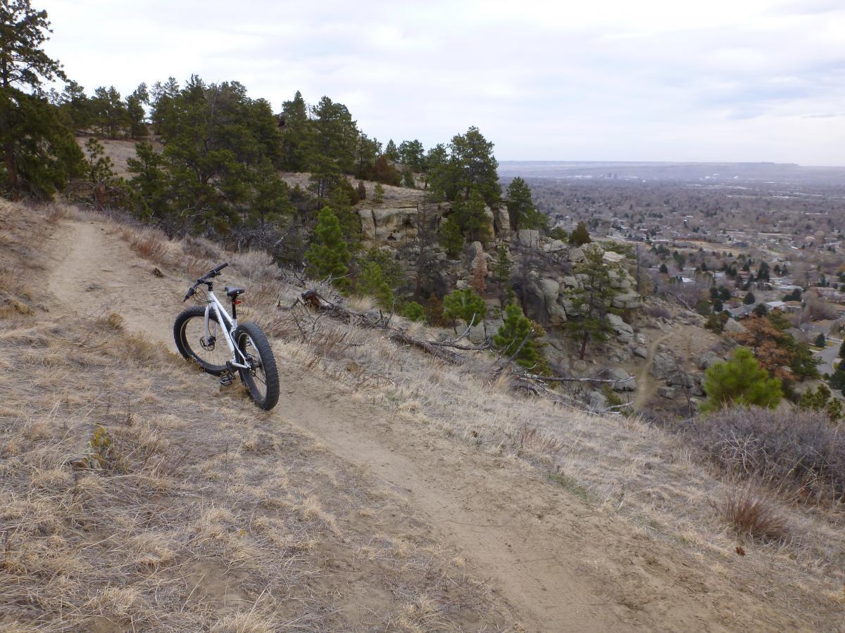 A mountain bike parked on a dirt trail overlooking a valley, surrounded by rocky outcrops and scattered pine trees. The sky is overcast, and a town can be seen in the distance. Zimmerman Trail mountain bike trail.