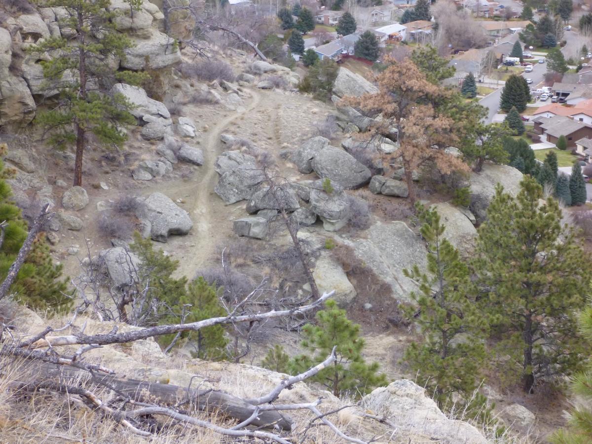 Alt text: A view of a rocky hillside with sparse vegetation, featuring scattered boulders and a dirt trail. In the background, a residential area is visible with houses and trees, surrounded by rolling hills. Zimmerman Trail mountain bike trail.