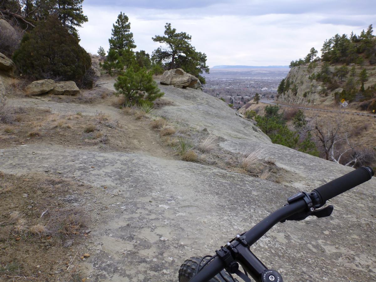 A mountain bike resting on rocky terrain, overlooking a valley with sparse vegetation and scattered trees. The landscape features a mix of dirt and rock, with a backdrop of rolling hills and cloudy skies. Zimmerman Trail mountain bike trail.
