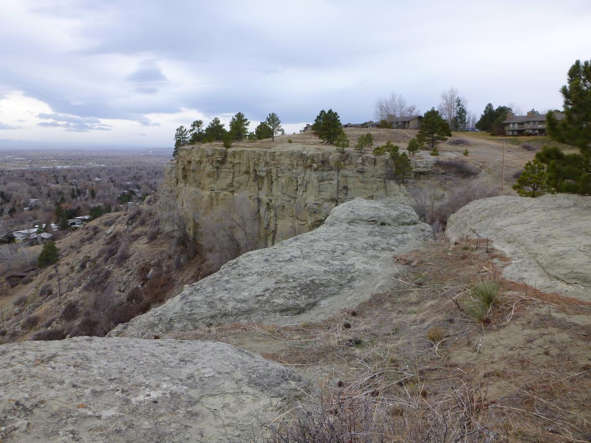 A rocky cliff with sparse vegetation overlooks a valley, under a cloudy sky. In the foreground, large boulders are visible, while in the background, a line of trees and distant houses can be seen along the hillside. The scene conveys a tranquil, natural landscape. Zimmerman Trail mountain bike trail.