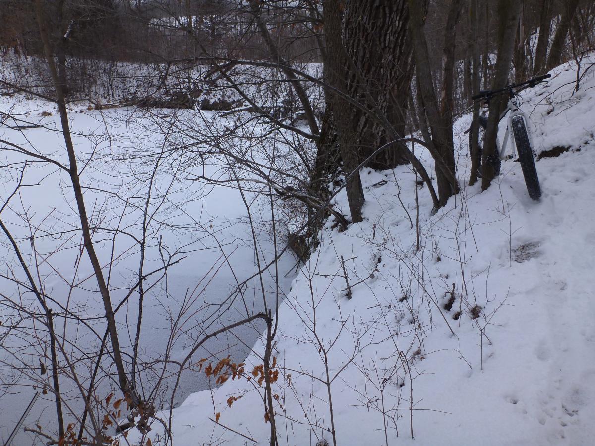 A winter scene featuring a frozen body of water surrounded by snowy terrain and bare trees. A bicycle is leaning against a tree on the right side, indicating a recreational area. The overall atmosphere is quiet and serene, illustrating a cold, snowy landscape. Mammoth Trail mountain bike trail.